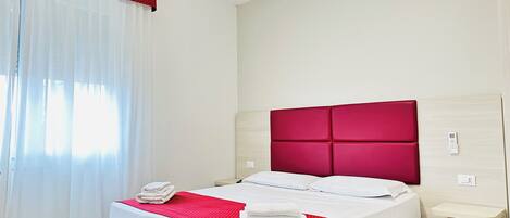 A hotel room with a bed, a bedside table, a red headboard, and a window with white curtains.