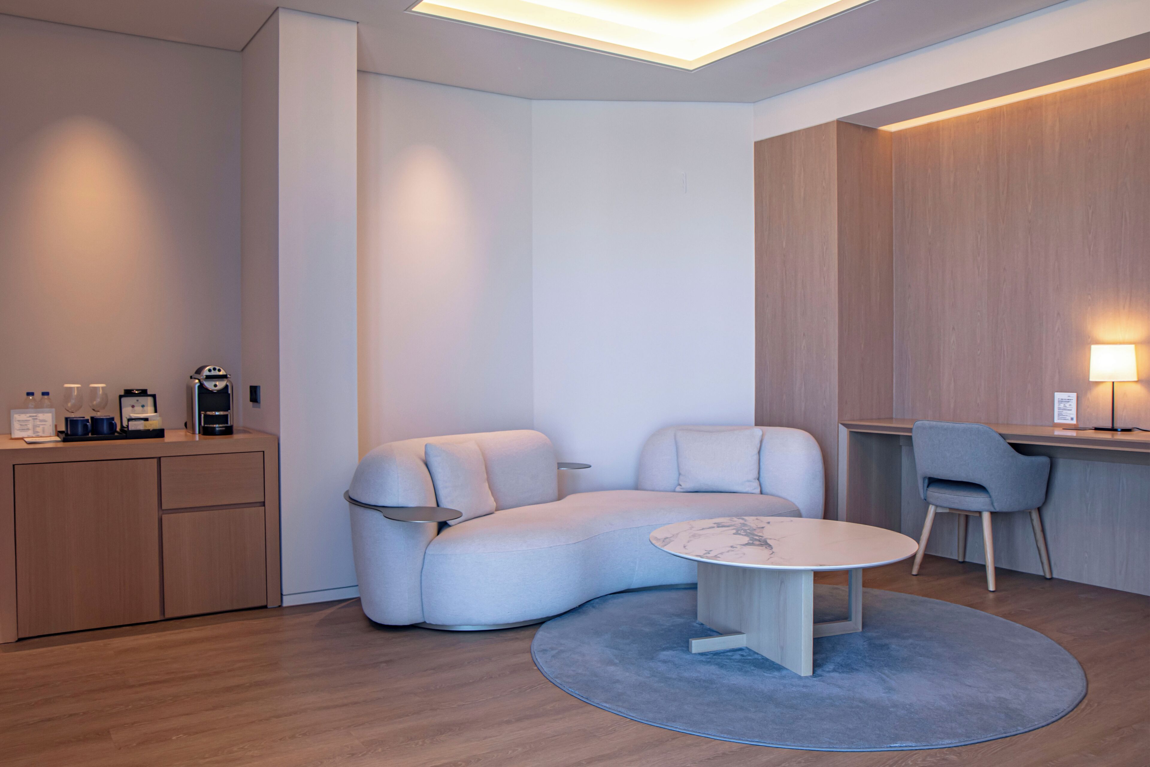Une pièce moderne et minimaliste, avec un canapé aux lignes courbes, une petite table ronde et un bureau avec une chaise. On y trouve également un meuble en bois sur lequel sont posés une machine à café et des bouteilles d’eau.