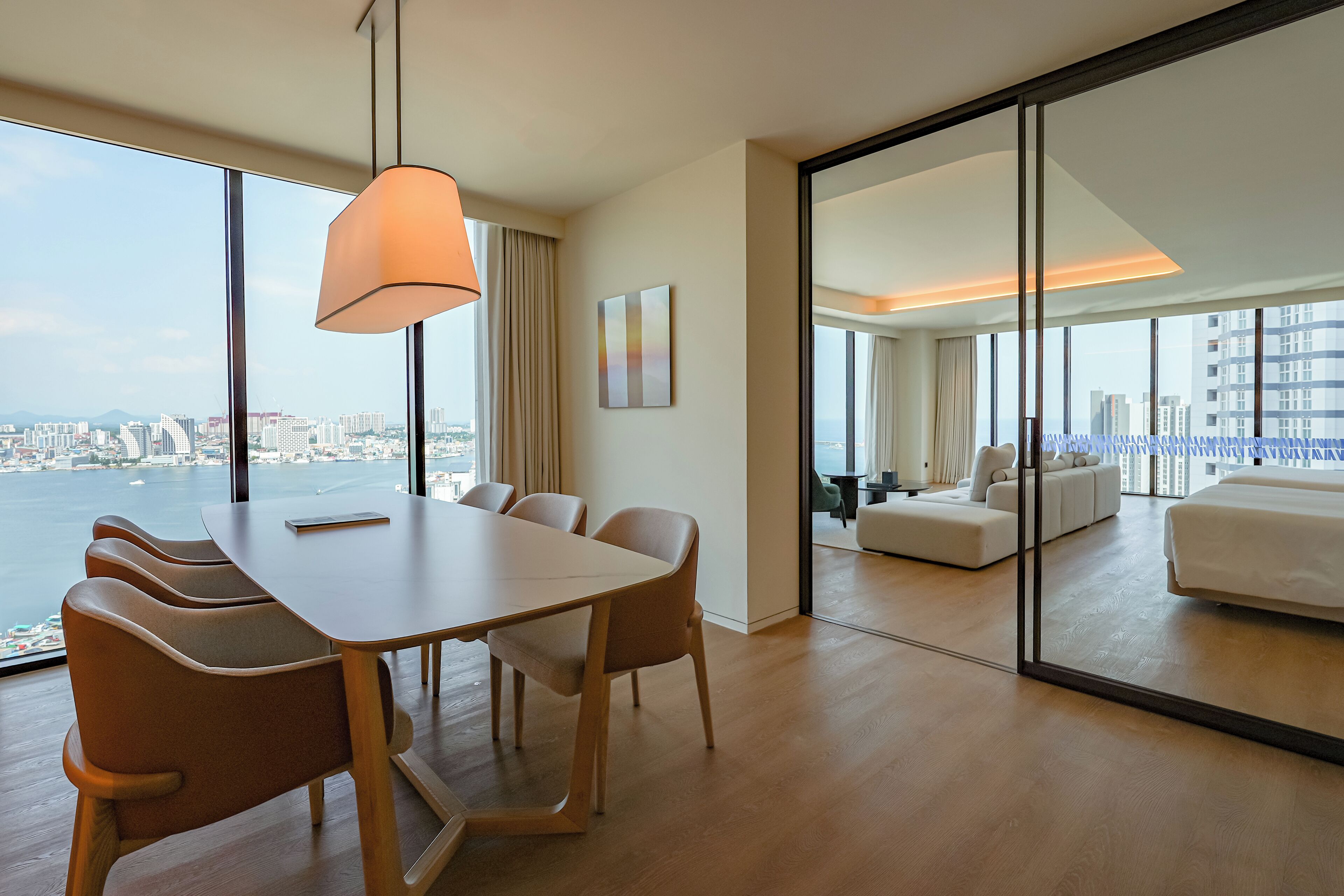 A modern hotel room with a dining area, a large window with a city view, and a separate living area with a sofa and a bed.