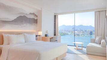 A hotel room with a large bed, a desk, a chair, and a sofa. There is a large window with a view of a city and mountains.