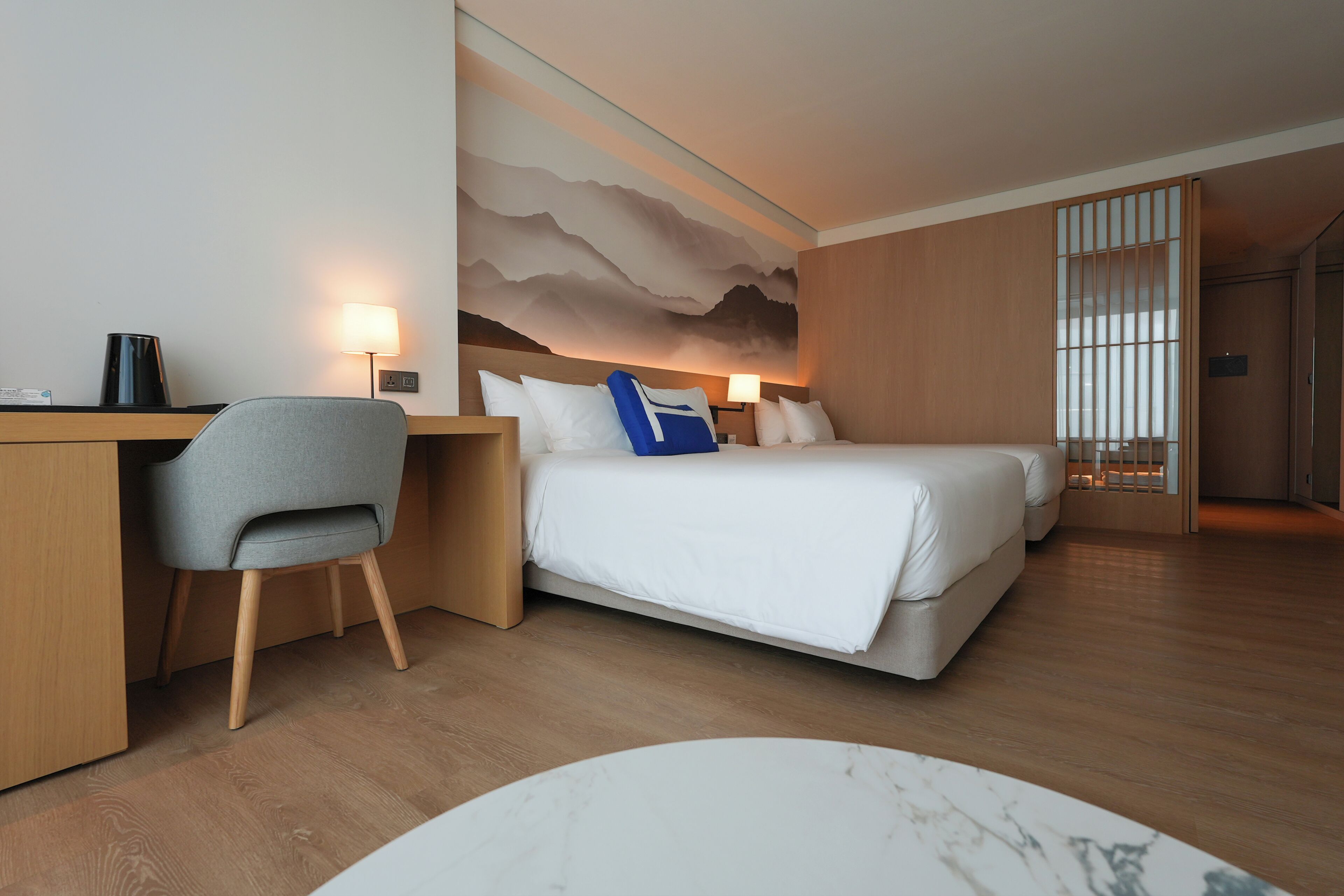 A hotel room with a desk, two beds, a chair, a lamp, and a wall mural.