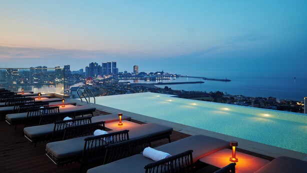Una piscina infinita con sillones de descanso y velas, con vistas a un paisaje urbano y al mar.