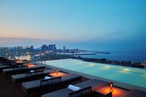 An infinity pool with lounge chairs and candles, overlooking a cityscape and the sea.