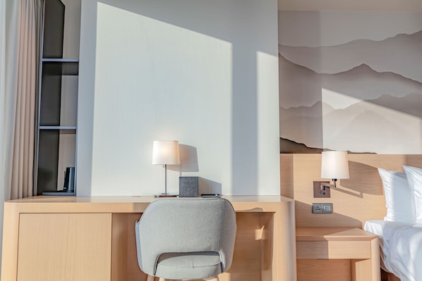 A modern hotel room with a wooden desk, a grey chair, a bed with white bedding, and a wall-mounted lamp.