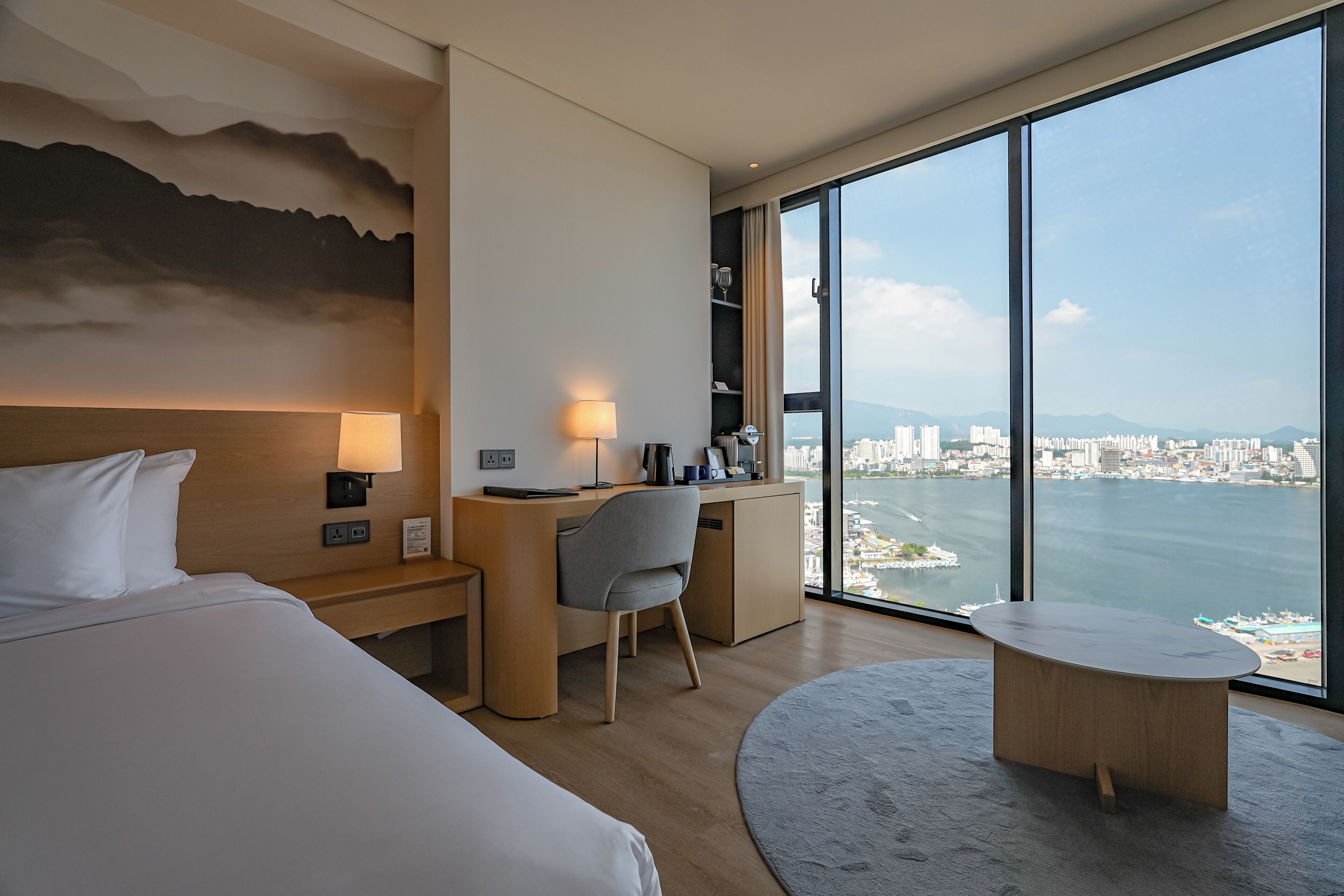 Una camera d'albergo con un letto, una scrivania, una sedia e un tavolo rotondo. Dispone di un'ampia finestra con vista sulla città e sul mare.