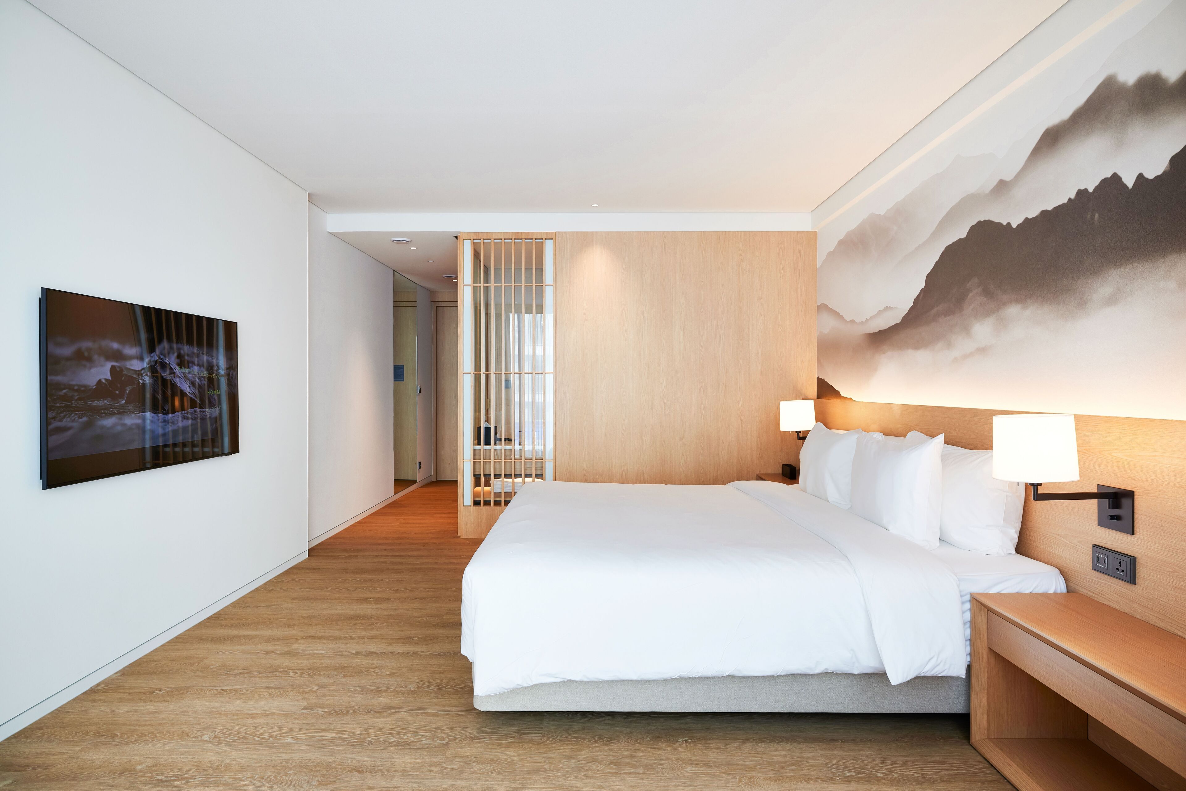 A modern hotel room with a large bed, a wooden headboard, a wall-mounted lamp, and a painting of mountains.