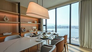 Un comedor moderno con una mesa grande dispuesta para una comida, ventanas amplias que ofrecen vistas a la ciudad y al agua, y una lámpara contemporánea sobre la mesa.
