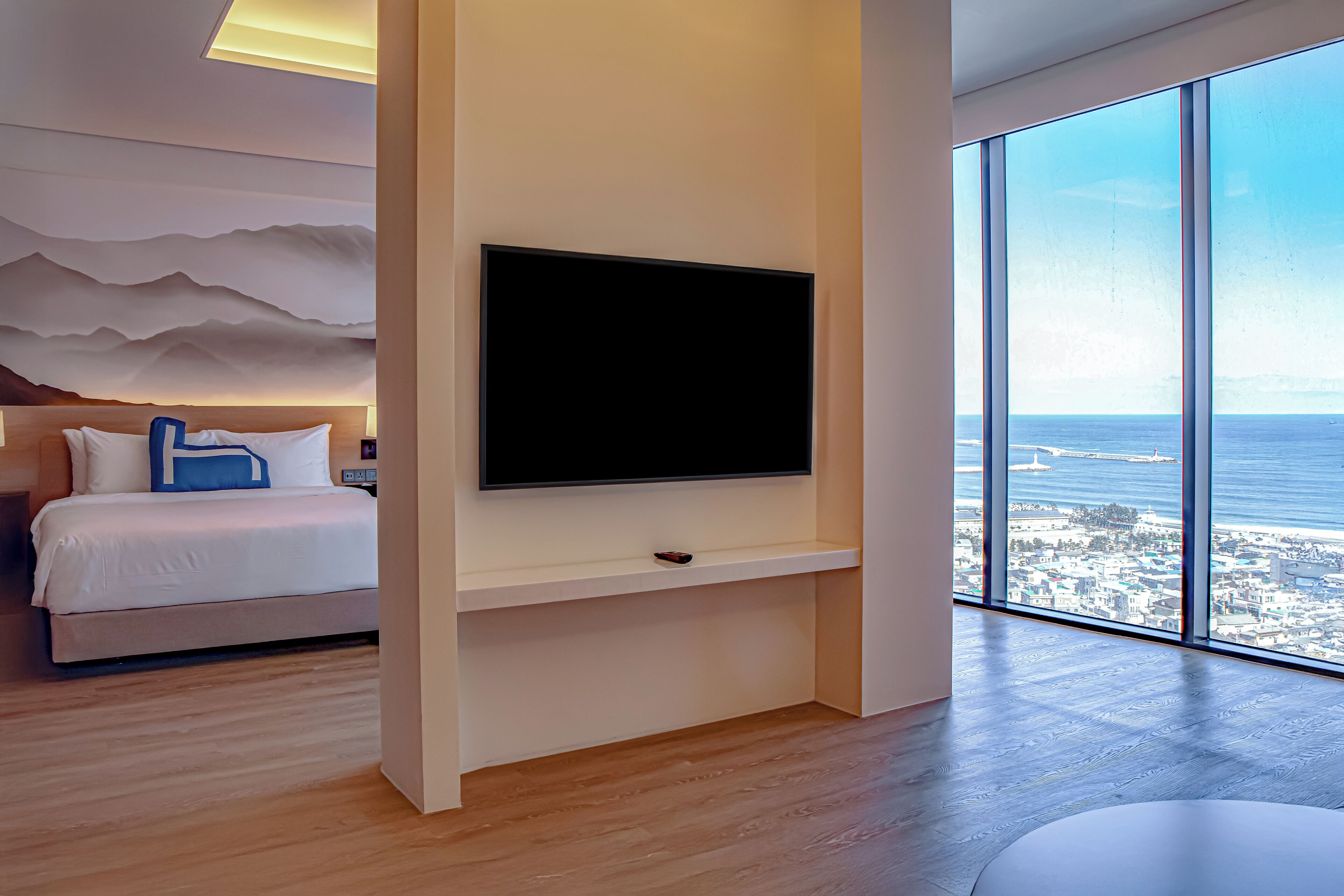 A modern hotel room with a large TV, a bed with white bedding, a wooden floor, and a view of the city and sea through large windows.
