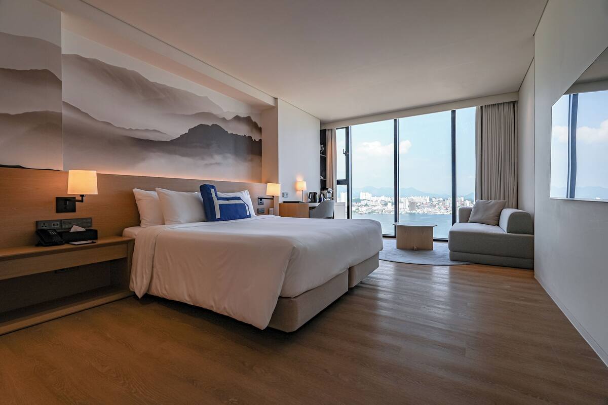 Ett modernt hotellrum med en stor säng, en soffa, ett litet soffbord och utsikt över staden genom stora fönster.