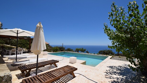 Uma área de piscina com espreguiçadeiras e um guarda-sol, com vista para um céu azul límpido e o oceano.