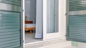 Een kamer met een bed, een klein tafeltje met een vaas en een fotolijstje, en een raam met een dunne gordijn.