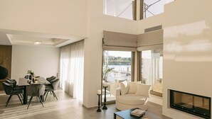 A spacious living area with a high ceiling, a modern chandelier, a fireplace, and large windows offering a view of the outdoors.