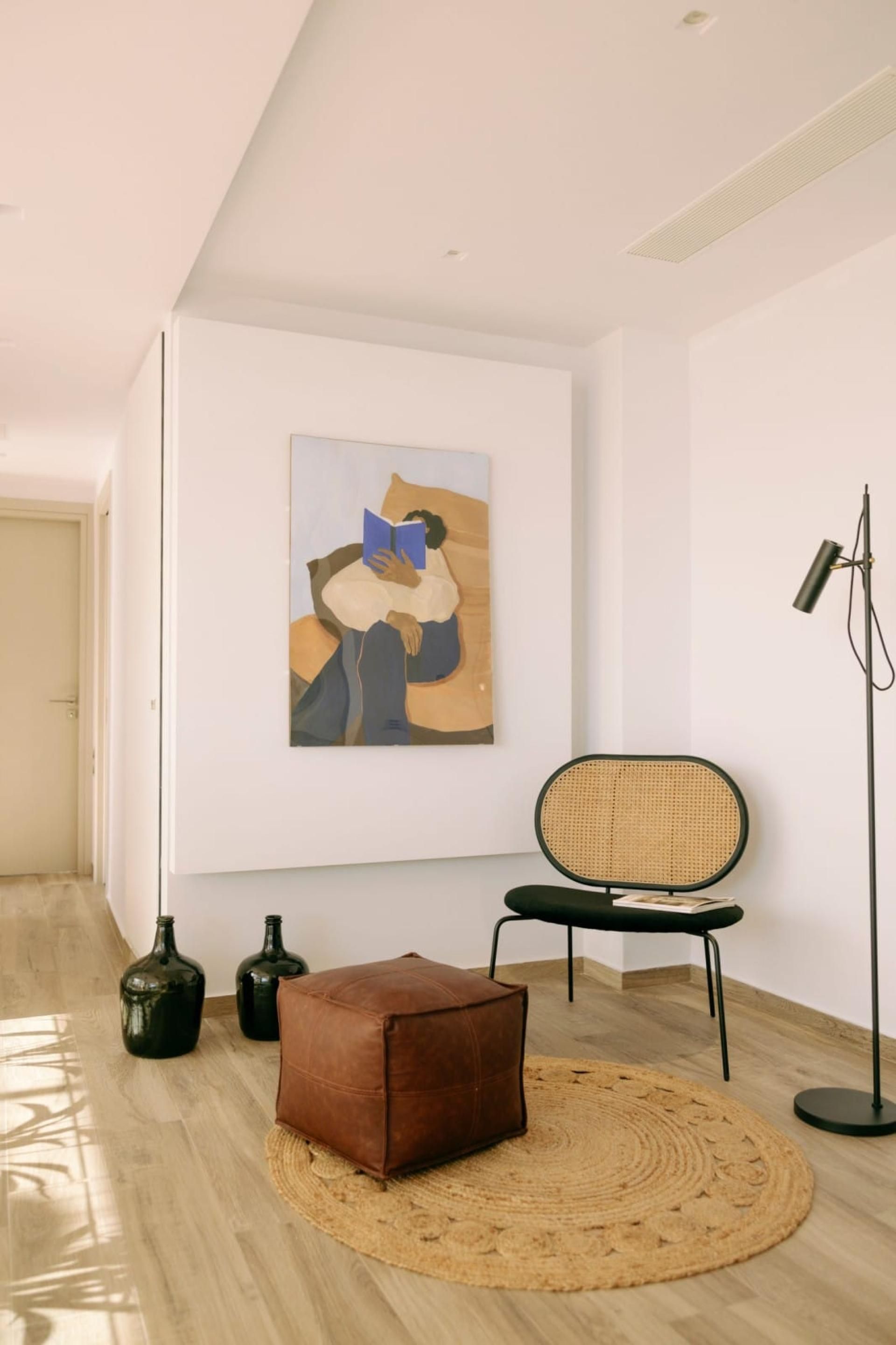 A modern interior with a wooden floor, a brown leather ottoman, a rattan chair, a floor lamp, and a painting on the wall.