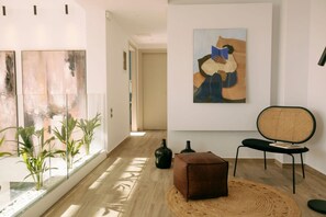 A room with a large window, a brown ottoman, a wicker chair, and a painting on the wall.