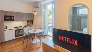 A modern kitchen with a dining area, a large TV screen displaying the Netflix logo, and a view of the outside through an open door. - Como Liberty Suites - Gatsby (Como)