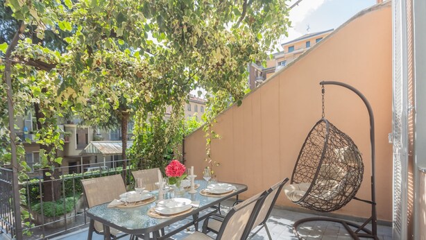 A dining area with a table set for four, surrounded by chairs, a hanging chair, and a potted plant with flowers. - Como Liberty Suites - Gatsby (Como)