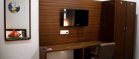 A hotel room with a wooden desk, a television mounted on the wall, a wardrobe, and a mirror with a decorative item.