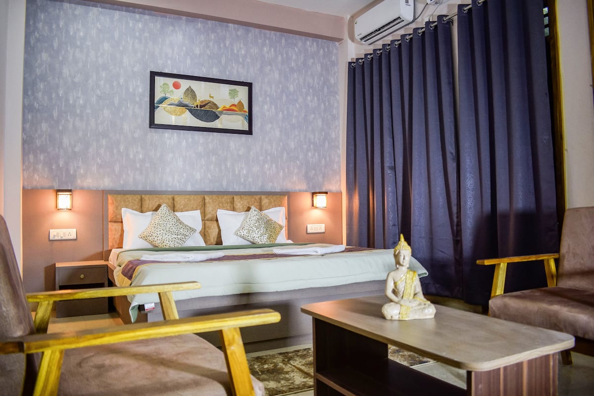 Quarto de hotel com cama, duas cadeiras, uma mesinha, ar condicionado fixado na parede e um quadro.