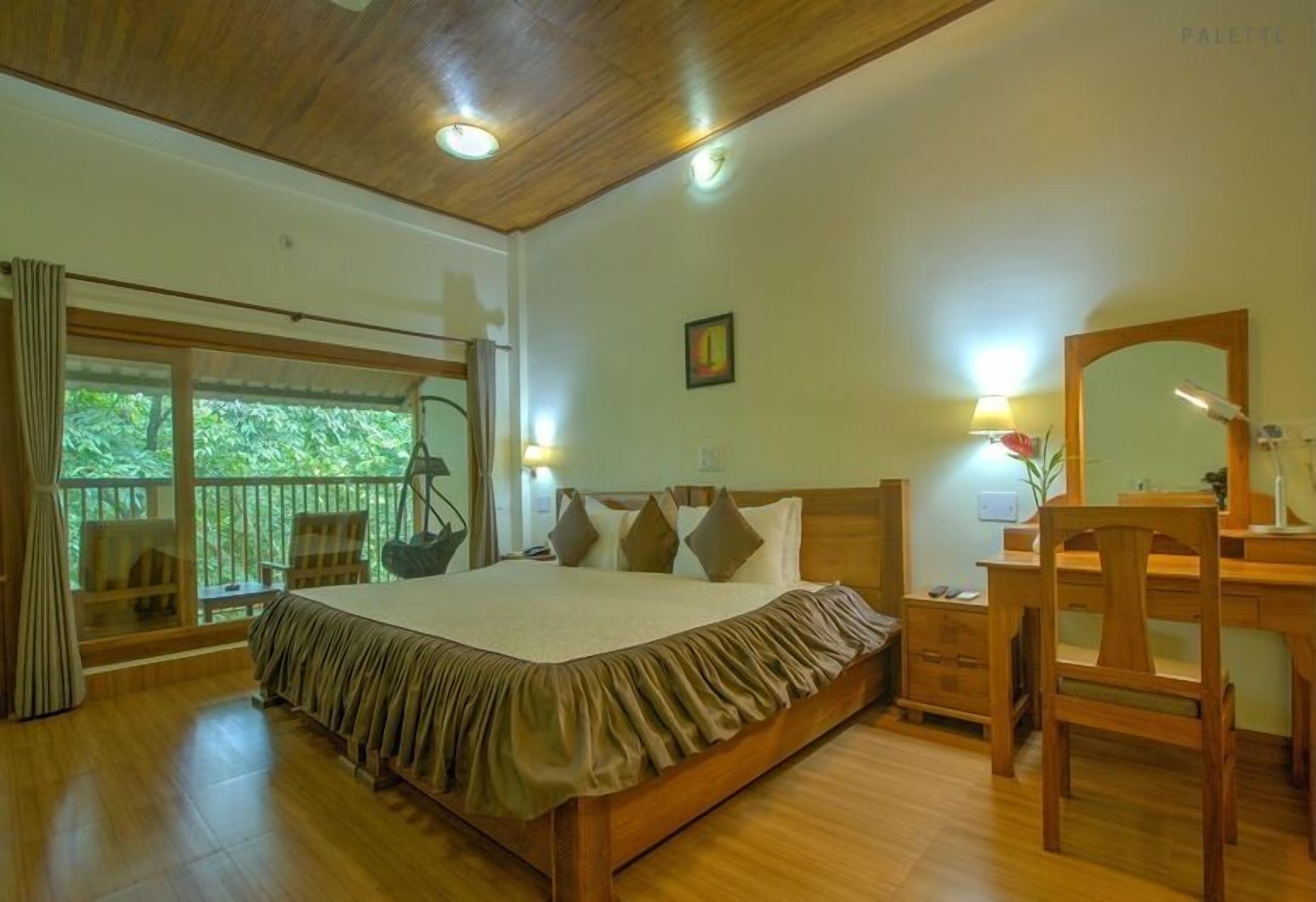 A bedroom with a large bed, wooden furniture, a desk, and a balcony with a view of greenery.