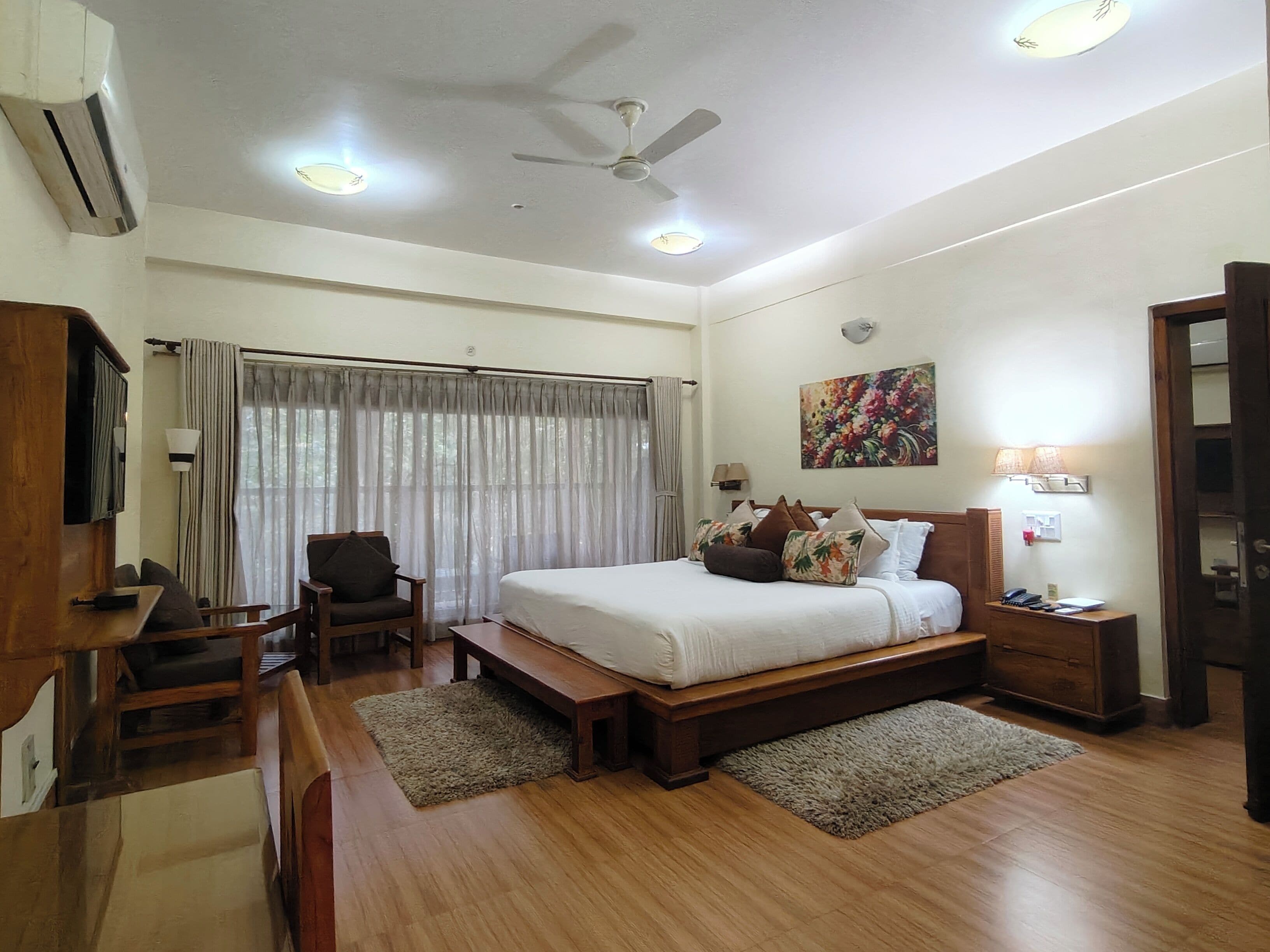 A hotel room with a large bed, a desk, a chair, a ceiling fan, and a painting on the wall.