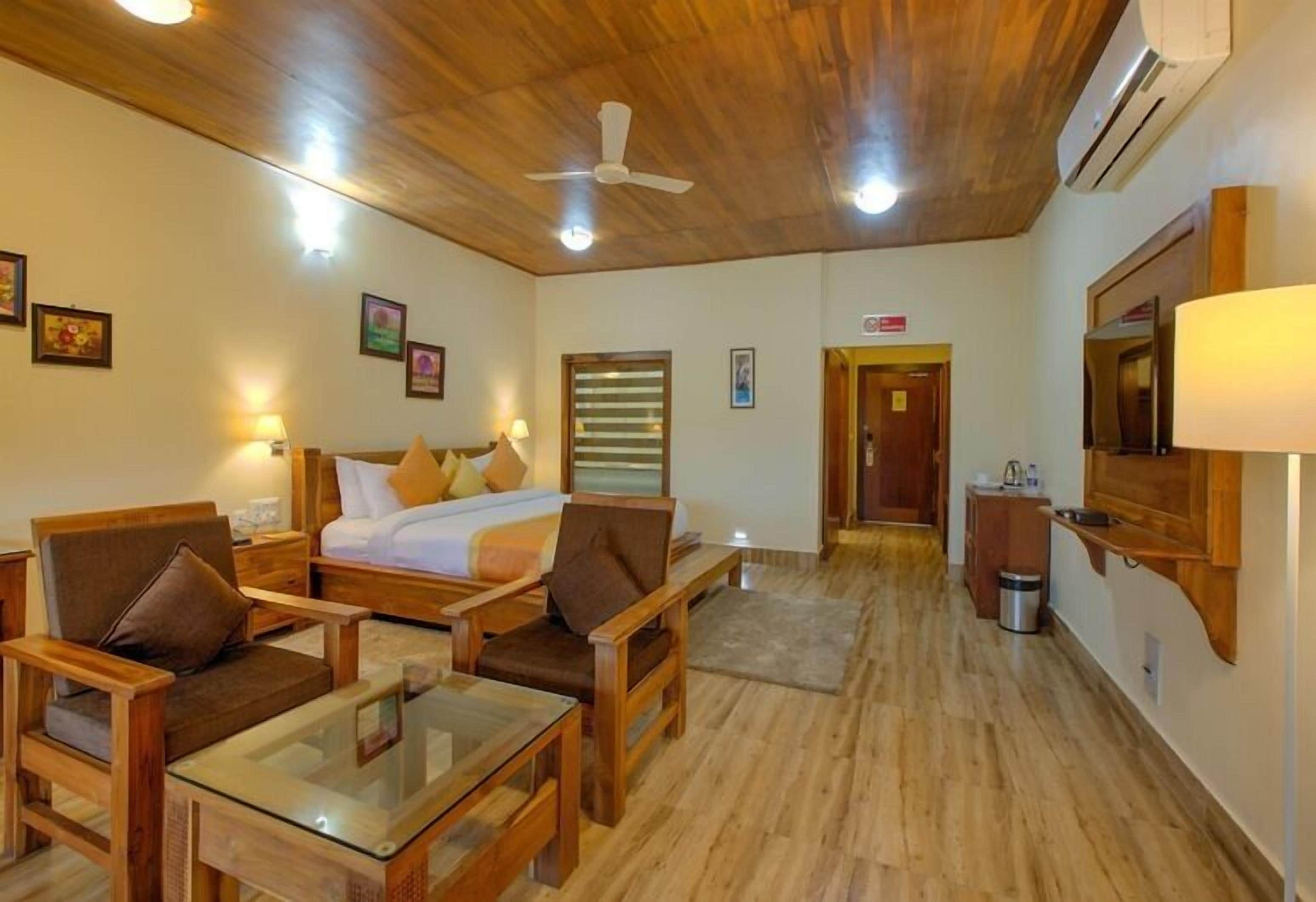 A hotel room with wooden flooring, a bed with pillows, a desk with a chair, a TV mounted on the wall, and a ceiling fan.