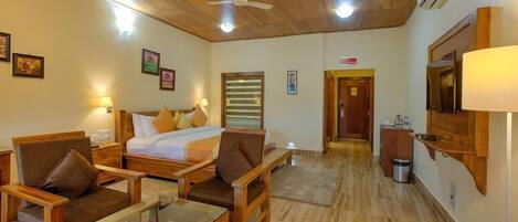 A hotel room with wooden flooring, a bed with pillows, a desk with a chair, a TV mounted on the wall, and a ceiling fan.