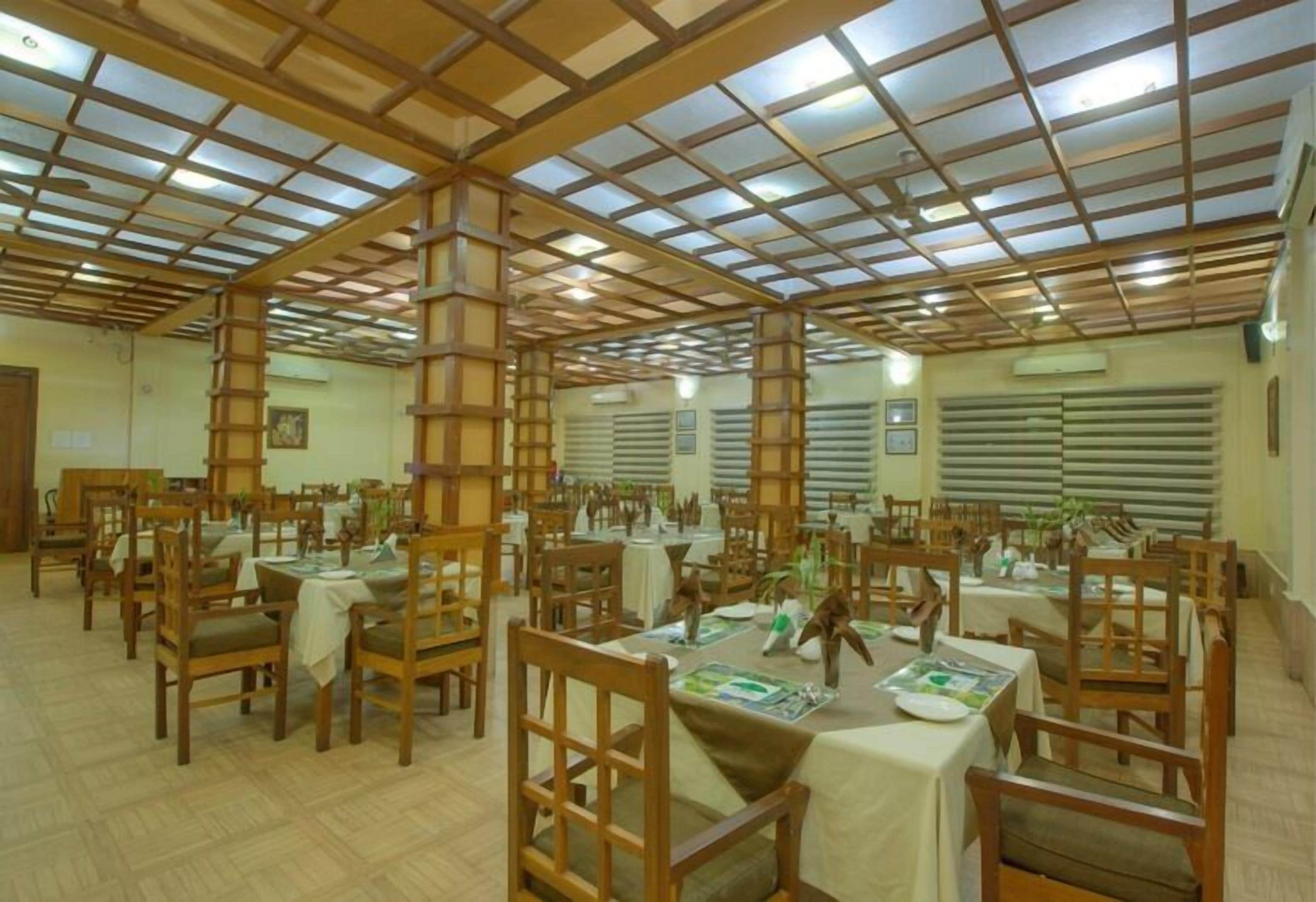 A dining area with wooden tables and chairs, a high ceiling with a grid pattern, and large windows with blinds.