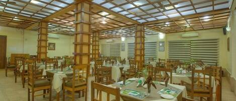A dining area with wooden tables and chairs, a high ceiling with a grid pattern, and large windows with blinds.
