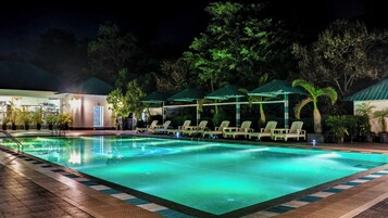 Una piscina con sillas de descanso y una zona de estar techada, rodeada de árboles y un edificio de fachada de ladrillo.