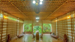 A traditional bamboo room with a central wooden altar, large windows, and a tiled floor. - Resort Borgos (Bokakhat)
