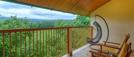 A wooden balcony with a view of lush greenery and mountains. There are two wooden chairs and a hanging chair.