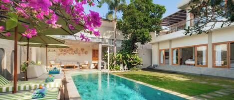 A swimming pool with lounge chairs and a covered seating area, surrounded by lush greenery and a modern building.