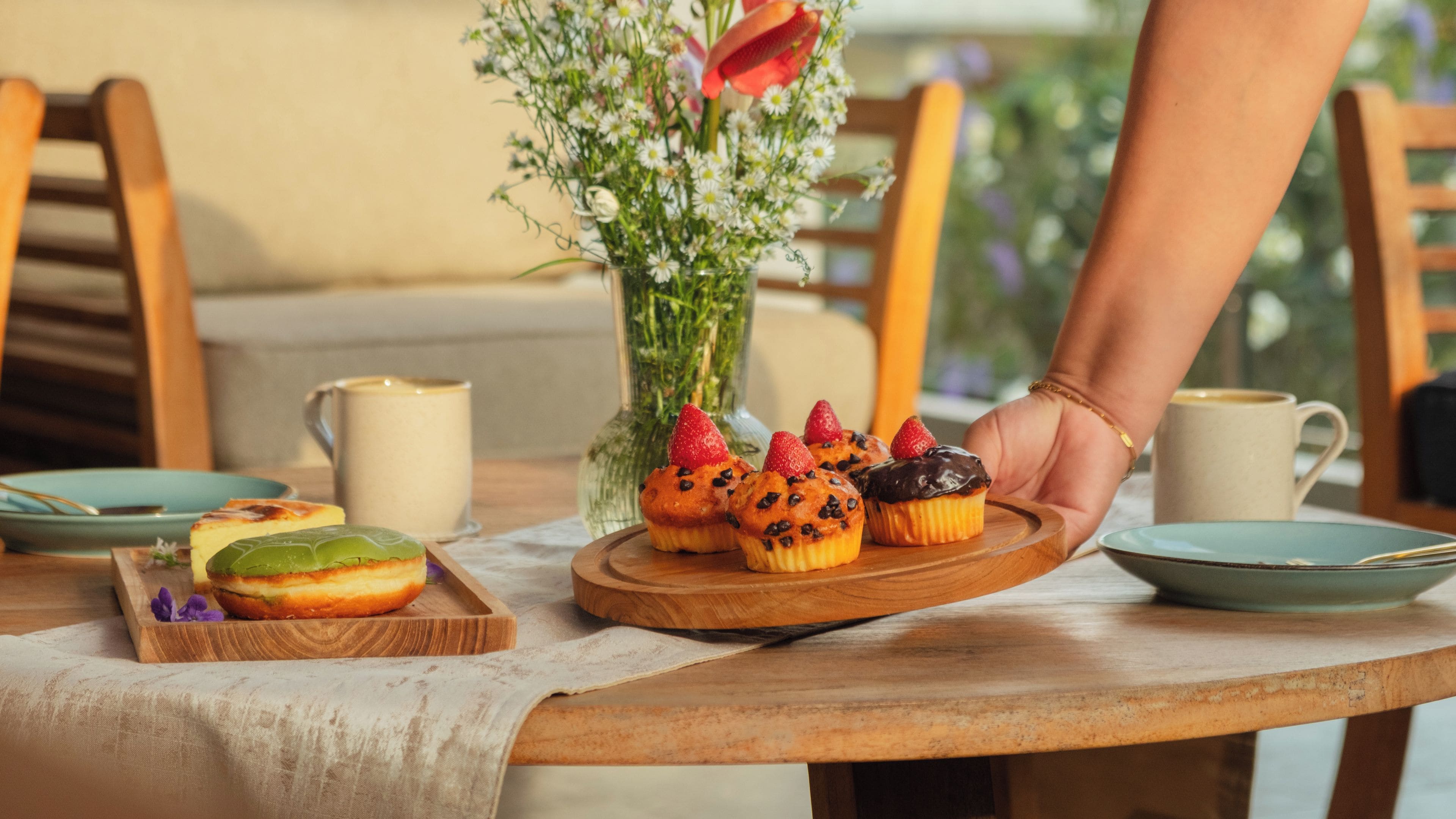 Una mesa de madera con una bandeja de pasteles, un florero con flores y una taza.