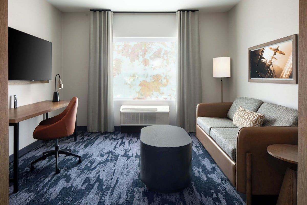 A hotel room with a desk, chair, sofa, ottoman, and a large window with curtains.