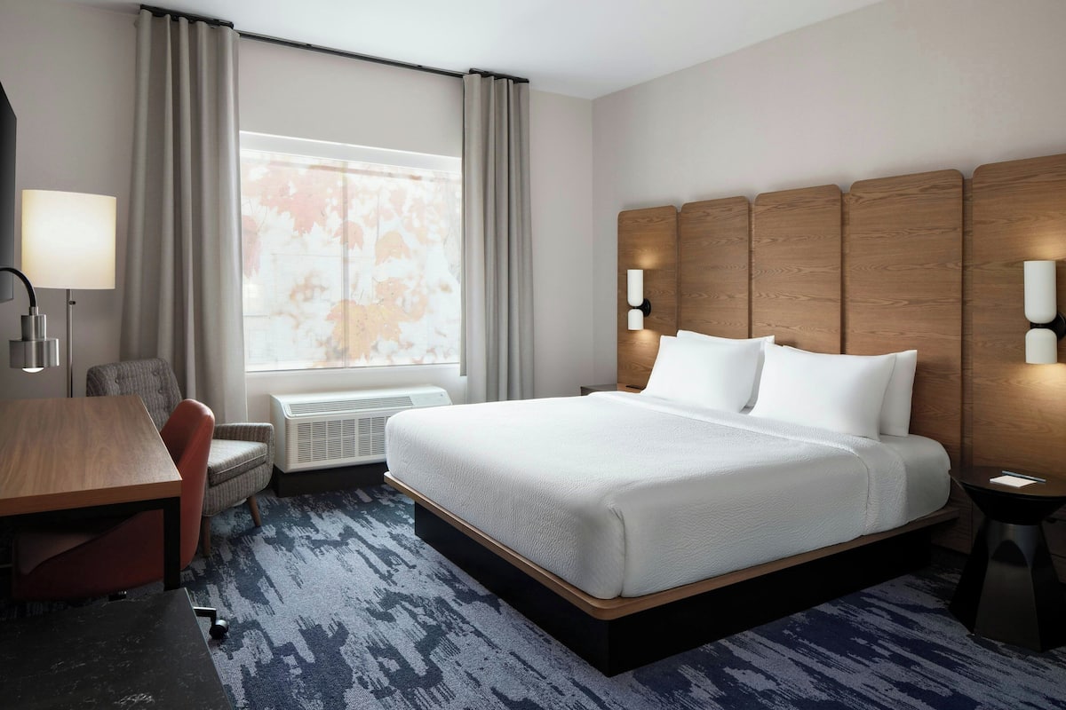 Habitación de hotel con una cama grande, un escritorio, una silla, un ventanal con cortinas e iluminación en la pared.