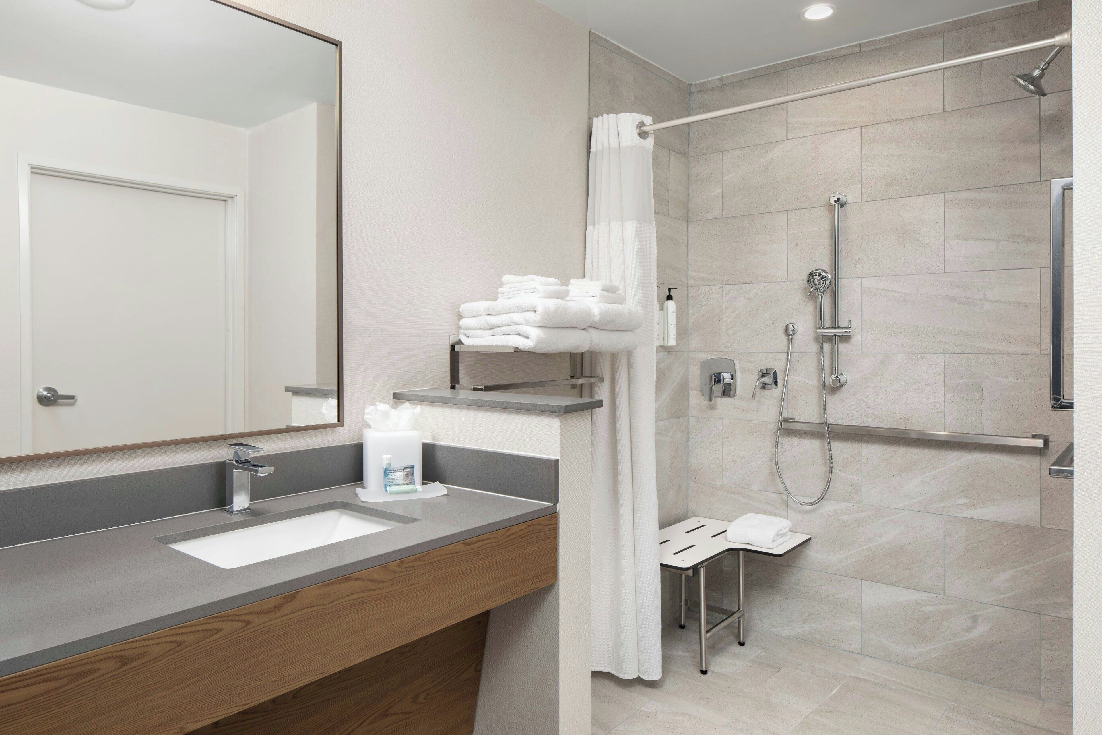 A modern bathroom with a large mirror, a sink with a wooden vanity, a shower with a glass door, and neatly stacked towels.