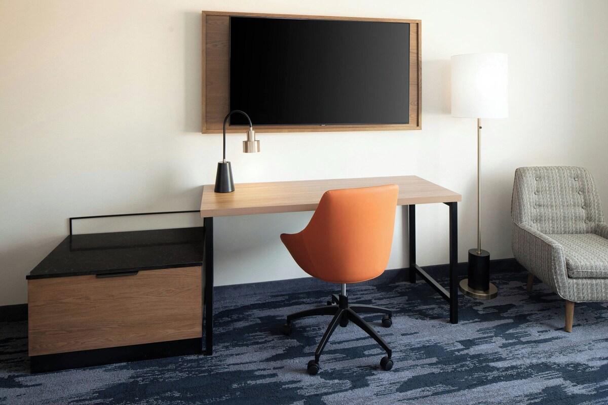 Una habitación de hotel con un escritorio de madera, una silla de oficina naranja, una lámpara negra, un sillón gris y un televisor de pantalla plana grande montado en la pared.