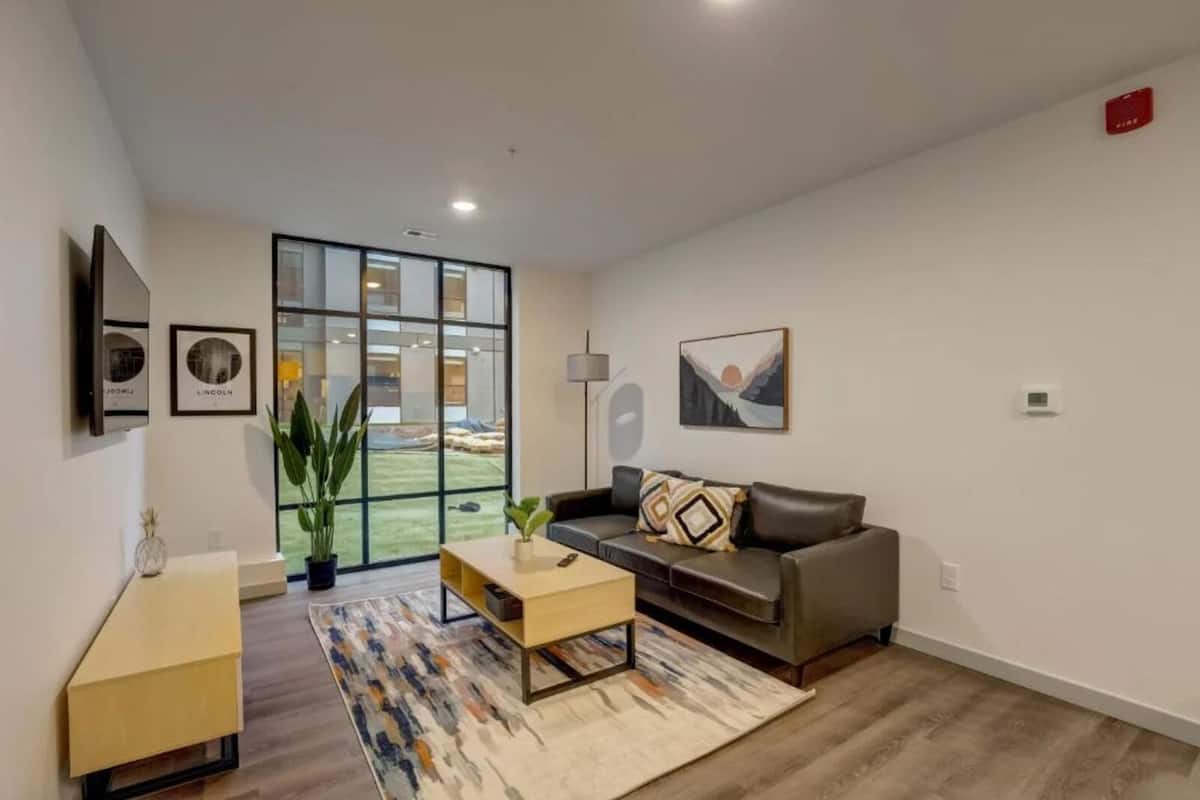 A modern living room with a grey sofa, a coffee table, and a large window offering a view of a green outdoor area.