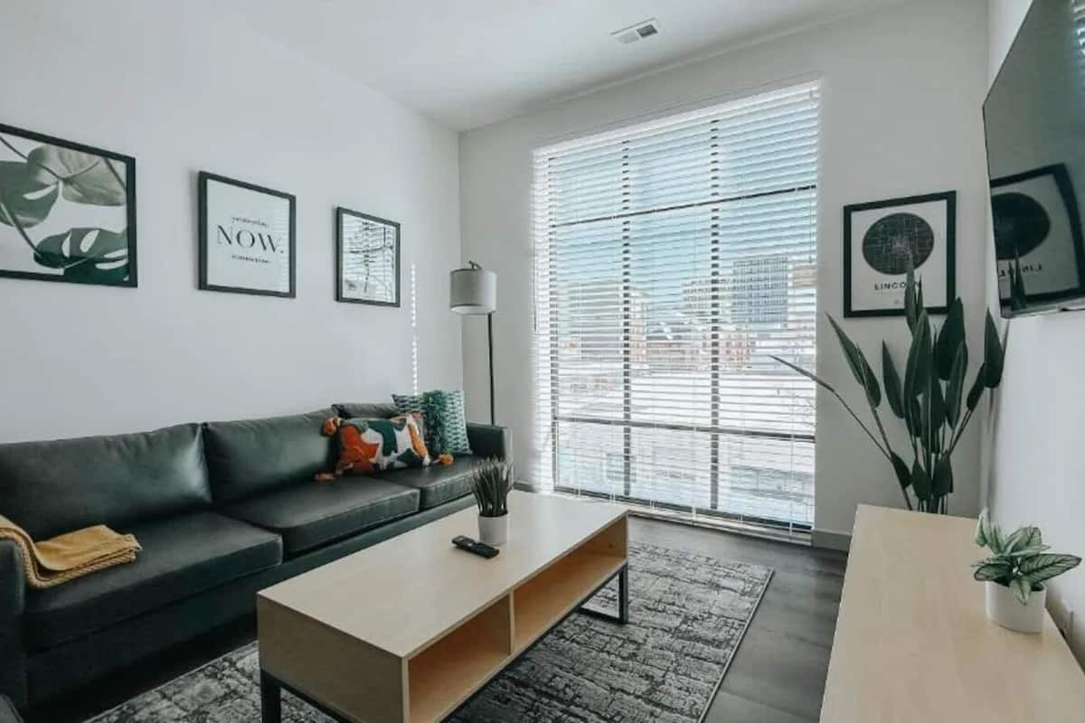 A modern living room with a black sofa, a wooden coffee table, and a large window with blinds.