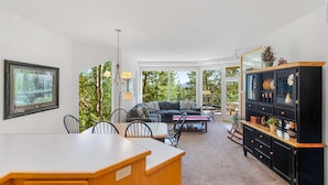A spacious living room with a large window, a dining area with a wooden table, and a black cabinet with decorative items. - Frosty Summit Condo at Mount Bachelor Village (Bend)