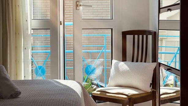 Un dormitorio con cama, una silla y una ventana que ofrece vistas a un edificio de ladrillo y estructuras metálicas azules.