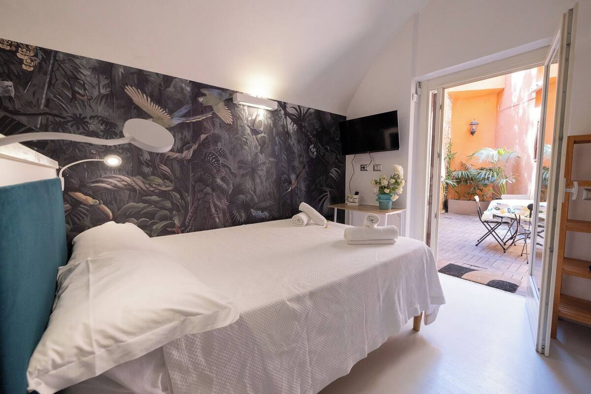 A bedroom with a bed, a nightstand, a wall-mounted TV, and a view of an outdoor patio with a table and chairs.