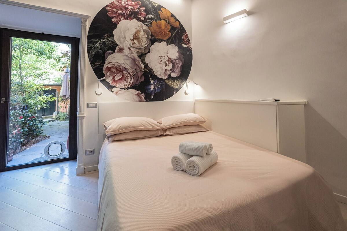 A bedroom with a bed, pillows, and a rolled towel. There is a circular floral artwork above the bed.