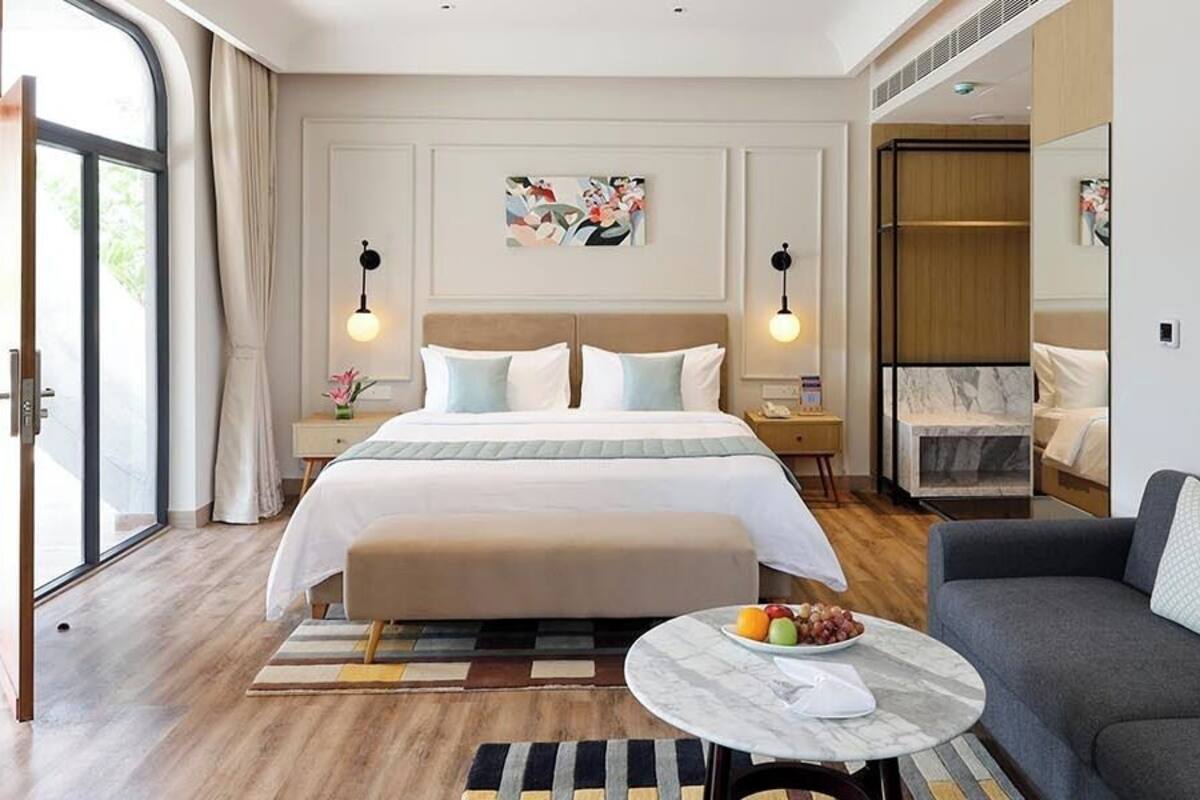 Una habitación de hotel moderna con una cama grande, un sofá, una mesita redonda con fruta y un ventanal amplio.