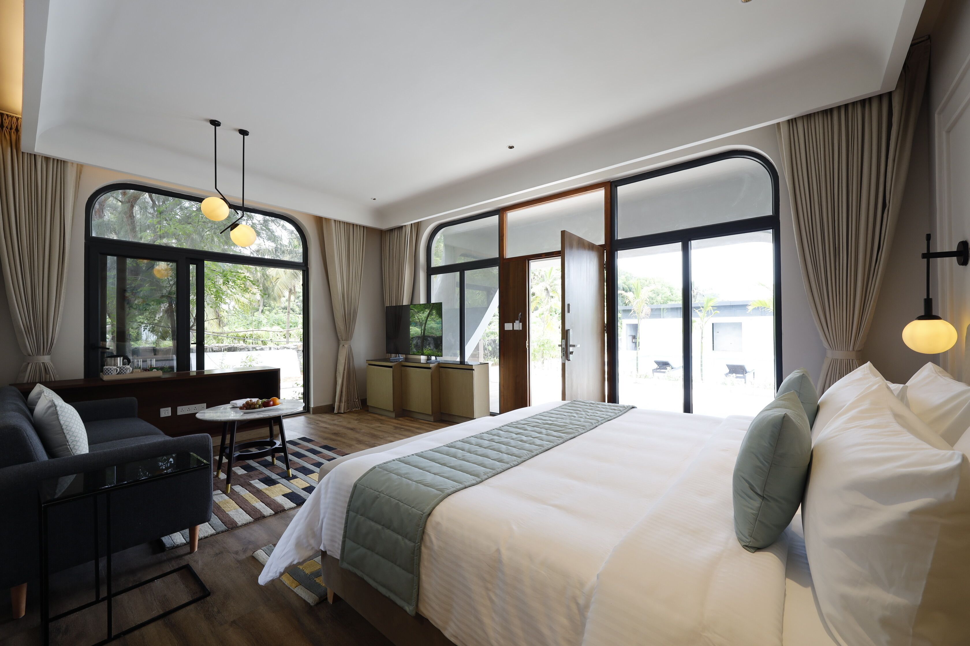 A modern hotel room with a large bed, a sofa, a small table, and a desk. There are large windows with curtains, a view of greenery outside, and a ceiling light.