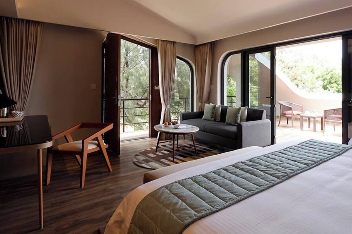 Habitación de hotel con una cama grande, un escritorio con una silla, un sofá y una mesita pequeña. Amplias ventanas con vistas al exterior.