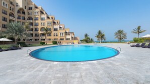 A swimming pool surrounded by lounge chairs and palm trees, with a building in the background. - Czechin - Kahraman Building (Ras Al Khaimah)