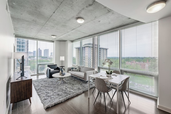 Una sala de estar moderna con sofá, mesa de centro y zona de comedor. Amplias ventanas ofrecen vistas a la ciudad.