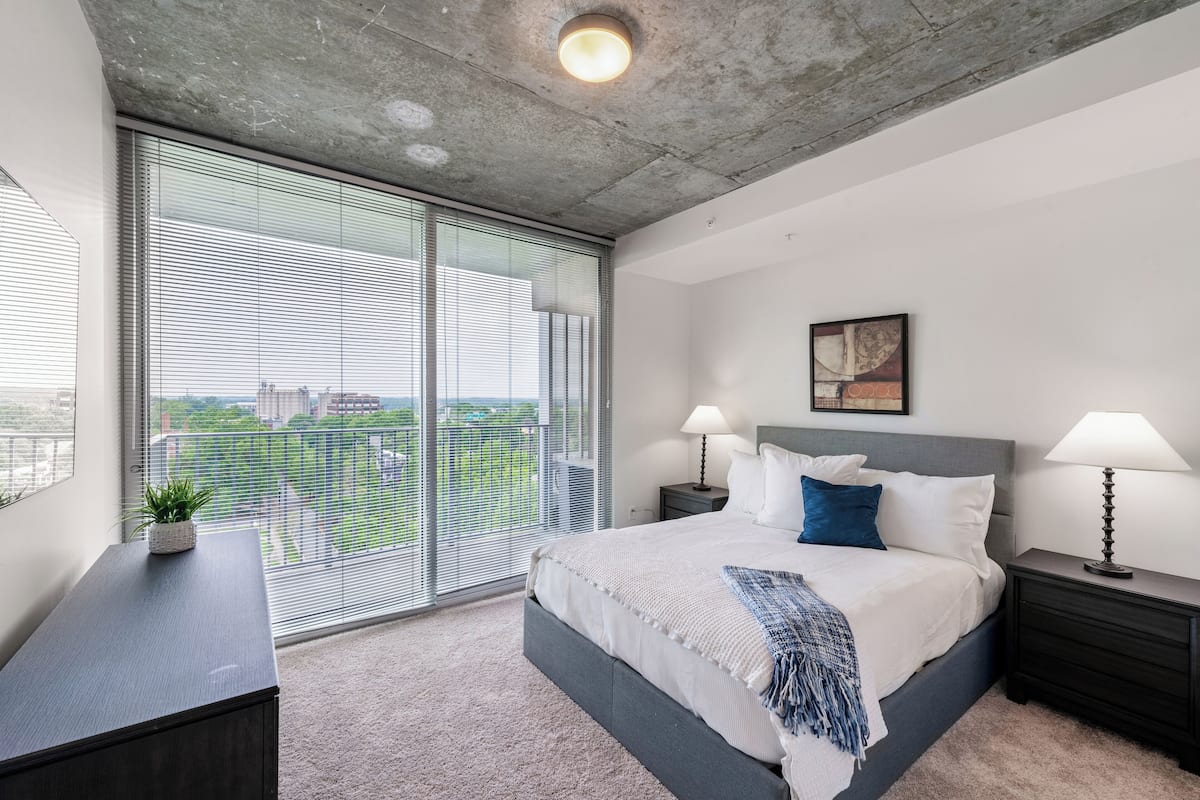 Une chambre moderne avec un grand lit, deux tables de chevet avec des lampes, une vue dégagée grâce à de grandes fenêtres du sol au plafond et un plafond en béton.