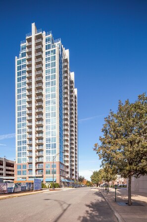 Un grand immeuble moderne, avec de nombreux balcons et de grandes fenêtres, se dresse dans un décor ensoleillé, avec une rue bordée d’arbres au premier plan.